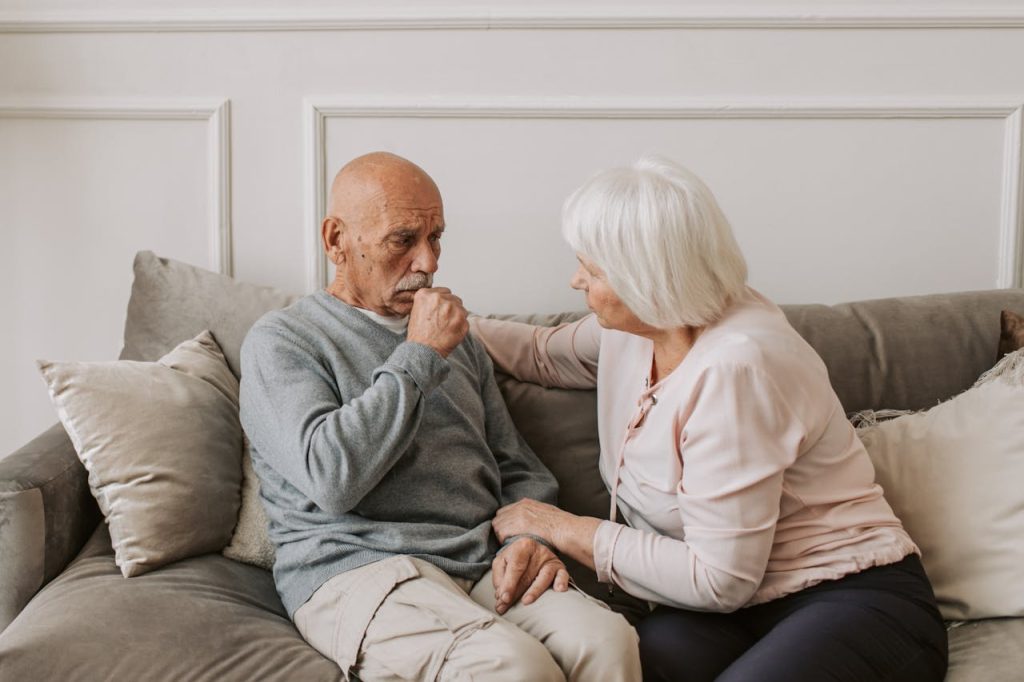 An elderly woman comforts a man coughing on a couch, showcasing care and affection.