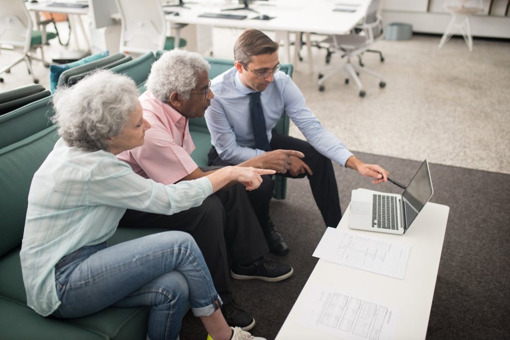 people-sitting-on-a-couch-while-looking-at-the-screen-of-laptop-8441823 Senior couple consulting with a professional on a laptop in a modern office setting.